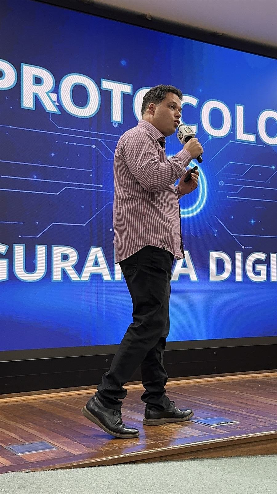 Elias Edenis caminhando no palco com o microfone, diante de um telão azul com as palavras “Protocolo” e “Segurança Digital”.